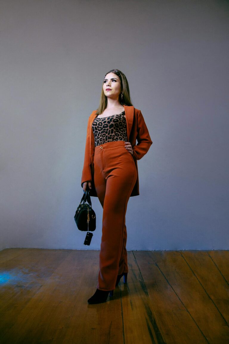 Fashionable woman poses confidently in a brown suit with leopard print top, holding a handbag indoors.